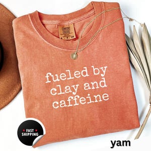 May include: A coral-colored t-shirt with the text "fueled by clay and caffeine" in white. A gold necklace with a pendant rests on the shirt. The shirt has a Comfort Colors tag and a "Fast Shipping" sticker.