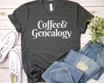 Może przedstawiać: Szary t-shirt z nadrukiem "Coffee & Genealogy" w kolorze białym na przodzie. Koszulka leży płasko na białym drewnianym tle z parą białych butów sportowych i niebieskimi dżinsami.