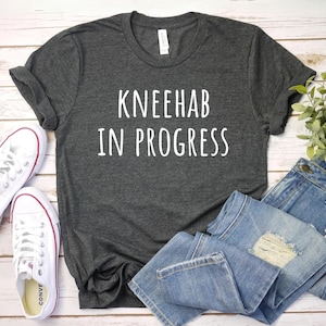 May include: A dark gray t-shirt with the text "Kneehab in Progress" printed in white. The shirt is paired with blue jeans and white sneakers.