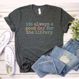 May include: Dark gray t-shirt with the text "it's always a good day for the library" in colorful letters. The shirt is paired with blue jeans and white sneakers with red trim, suggesting a casual, everyday style.