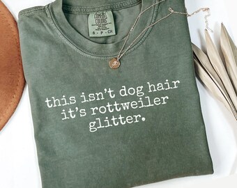 May include: A moss green t-shirt with the text "this isn't dog hair it's rottweiler glitter" in white. A gold necklace with a pendant rests on the shirt. The shirt is labeled "Comfort Colors" and is size S-P-CH.