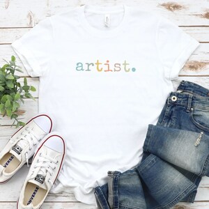 Peut inclure: T-shirt blanche avec le mot "artist." imprim&eacute; en couleurs arc-en-ciel sur le devant. Le t-shirt est pos&eacute; sur une surface en bois blanc avec une paire de baskets blanches et un jean bleu.