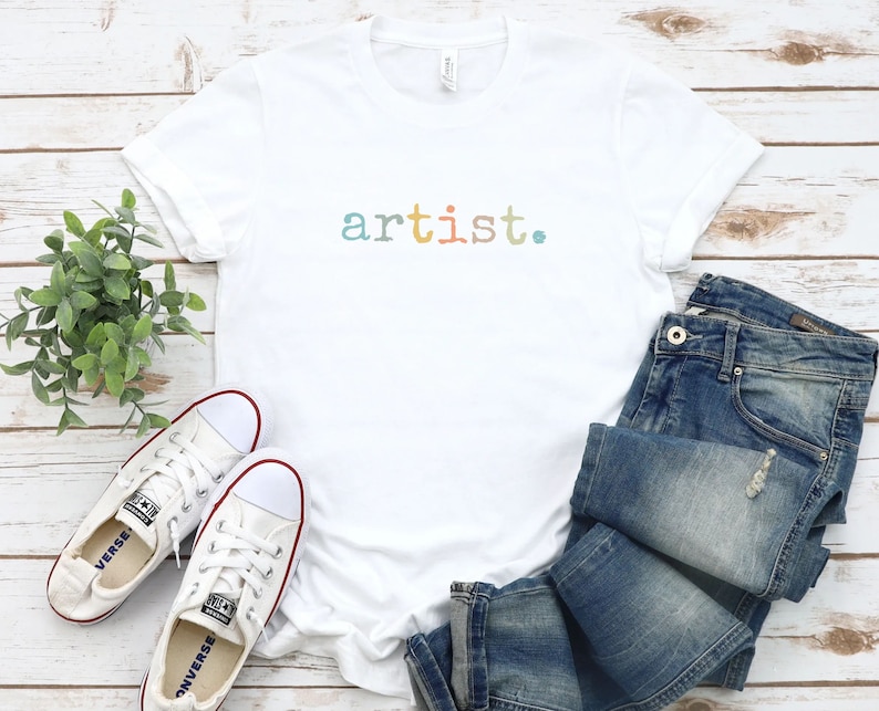 Peut inclure: T-shirt blanche avec le mot "artist." imprim&eacute; en couleurs arc-en-ciel sur le devant. Le t-shirt est pos&eacute; sur une surface en bois blanc avec une paire de baskets blanches et un jean bleu.