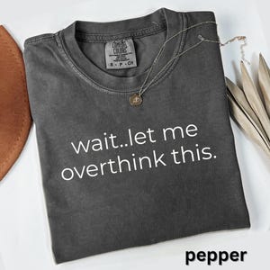 May include: A dark gray Comfort Colors t-shirt with the text "wait..let me overthink this." A silver necklace with a small pendant rests on the shirt. The word "pepper" is at the bottom of the shirt.