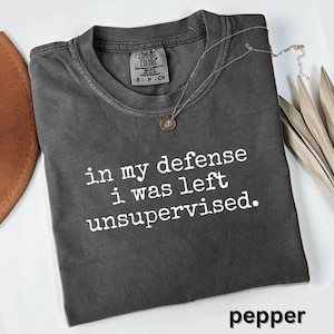 May include: A dark gray t-shirt with the text "in my defense i was left unsupervised." The shirt has a crew neck and a small tag on the upper left. A silver necklace with a pendant is draped across the shirt. The word "pepper" is at the bottom.