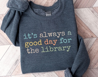 May include: A dark heather gray crewneck sweatshirt featuring the text "it's always a good day for the library" printed in multiple colors including light blue, yellow, orange, and light green across the center of the chest area.
