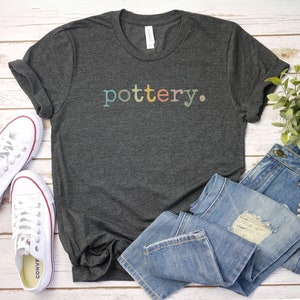 May include: A dark gray t-shirt with the word "pottery" in colorful letters. The shirt is paired with blue jeans and white canvas sneakers with red accents. A small green plant is visible in the background.