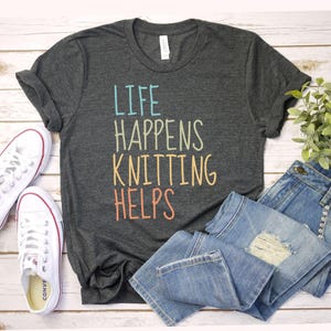 May include: Dark gray t-shirt with the text "LIFE HAPPENS KNITTING HELPS" in colorful letters. The shirt is paired with blue jeans and white sneakers with red accents. A sprig of greenery adds a touch of freshness.