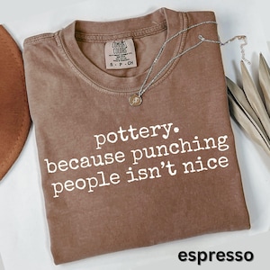 May include: Espresso-colored t-shirt with the text "pottery. because punching people isn't nice" in white. A silver necklace with a small pendant rests on the shirt. The shirt has a Comfort Colors label.