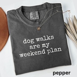 May include: A dark gray t-shirt with the text "dog walks are my weekend plan" in white. A silver necklace with a pendant is on the shirt. The word "pepper" is at the bottom of the shirt.