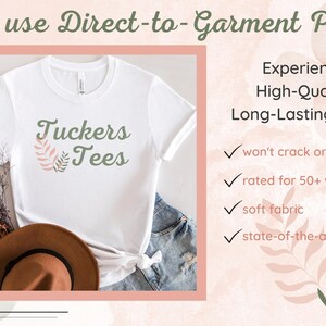 Peut inclure: Un t-shirt blanc avec un motif floral vert et rose et le texte "Tuckers Tees". L'image montre le t-shirt sur une table avec un chapeau marron et un jean bleu. Le texte "We use Direct-to-Garment Print" est en haut de l'image. Le texte "Experience High-Quality, Long-Lasting Prints" est &agrave; droite de l'image. Sous le texte, il y a quatre coches avec le texte suivant : "won't crack or peel", "rated for 50+ washings", "soft fabric", et "state-of-the-art print".