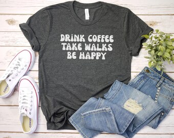 May include: A dark gray t-shirt with the text "Drink Coffee Take Walks Be Happy" in white, retro-style lettering. The shirt is laid flat on a white wooden surface with a pair of white sneakers and blue jeans.