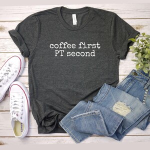 May include: Dark gray t-shirt with the text "coffee first PT second" in white. The shirt is paired with light blue denim jeans and white canvas sneakers with red accents. A sprig of greenery is visible.