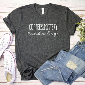 May include: A dark gray t-shirt with the text "Coffee & Pottery kinda day" in white lettering. The shirt is laid out on a white wooden surface with a pair of white sneakers and blue jeans.