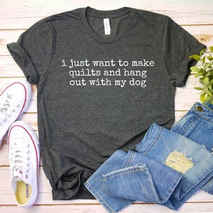 May include: A dark grey t-shirt with the text "i just want to make quilts and hang out with my dog" printed in white. The shirt is shown on a wooden surface with a pair of white sneakers and a pair of blue jeans.