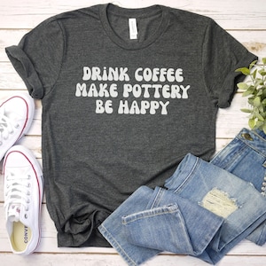 May include: A dark gray t-shirt with the text "Drink Coffee Make Pottery Be Happy" in white, retro-style lettering. The shirt is laid out on a white wooden surface with a pair of white sneakers and blue jeans.