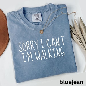 May include: A bluejean-colored Comfort Colors t-shirt with the text "SORRY I CAN'T I'M WALKING" in white. A gold-colored necklace with a pendant is draped across the shirt. The shirt is size small.