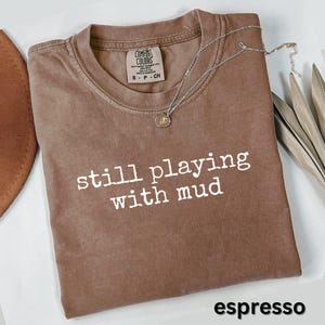May include: Espresso-colored t-shirt with the phrase "still playing with mud" in white lettering. A silver necklace with a pendant rests on the shirt. The shirt has a crew neck and a tag that says "Comfort Colors".