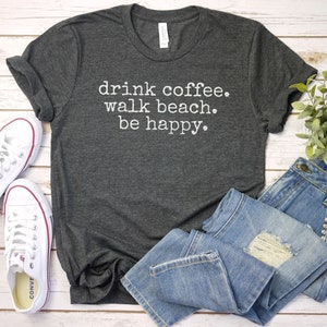 May include: A dark gray t-shirt with the text "drink coffee. walk beach. be happy." printed in white. The shirt is laid flat on a wooden surface with a pair of white sneakers and blue jeans.