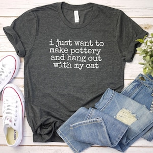 May include: A dark gray t-shirt with the text "i just want to make pottery and hang out with my cat" printed in white.