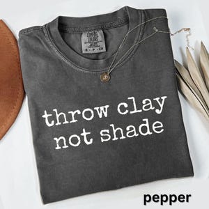 May include: A dark gray t-shirt with the text "throw clay not shade" in white. The shirt has a crew neck and a small tag at the neckline. A silver necklace with a pendant is draped on the shirt. The word "pepper" is at the bottom.
