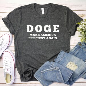 May include: A dark gray t-shirt with the text "DOGE MAKE AMERICA EFFICIENT AGAIN" in white letters. The shirt is laid out on a wooden surface with a pair of white sneakers and blue jeans.