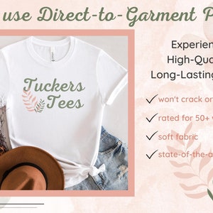 May include: White t-shirt with a green and pink floral design and the text "Tuckers Tees". The image shows the t-shirt on a table with a brown hat and blue jeans. The text "We use Direct-to-Garment Print" is at the top of the image. The text "Experience High-Quality, Long-Lasting Prints" is on the right side of the image. Below the text are four check marks with the following text: "won't crack or peel", "rated for 50+ washings", "soft fabric", and "state-of-the-art print".