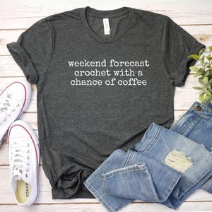 A gray t-shirt with white text, a pair of white sneakers, and a pair of blue jeans are arranged on a wooden surface.