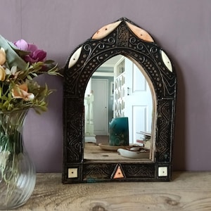 May include: An arched wall mirror with a dark, intricately carved wooden frame featuring inlaid light-coloured bone or wood accents in geometric and leaf shapes. The frame displays detailed scrollwork patterns and a pointed top design.