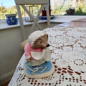 May include: A ceramic figurine of a hedgehog wearing a pink dress, white apron, and bonnet. The hedgehog is standing on a blue base with knitting needles. The figurine is on a white lace tablecloth.