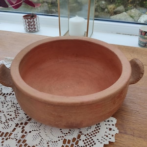 May include: A terracotta bowl, reddish-brown in colour, with a wide, shallow design and two small handles. The bowl is unglazed and sits on a white lace tablecloth. It is likely used for cooking or serving food. The bowl's diameter is approximately 30 cm.