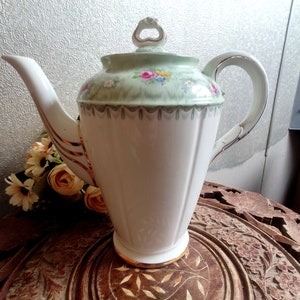 Peut inclure: Théière en porcelaine blanche avec couvercle vert clair et motif floral. La théière a une bordure dorée sur la poignée, le bec et la base. Posée sur une table en bois.