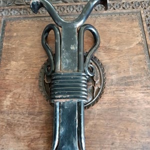 May include: A black metal wall hook featuring a central vertical bar with a flared base and a top section that branches into two curved arms. The middle of the bar is wrapped with a series of horizontal metal coils, creating a decorative accent.