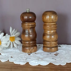 May include: Two wooden salt and pepper grinders with a polished brown finish and a rounded, tiered shape. The left grinder features a small metallic knob at the top, while the right grinder has a smooth, rounded top. Both display natural wood grain.