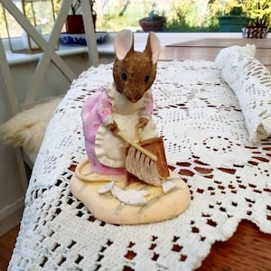 Può includere: Statuetta dettagliata di un topo con un vestito rosa e un grembiule bianco, che tiene una scopa. Il topo è in piedi su una base beige con piccoli pezzi di carta. La statuetta è posta su una tovaglia di pizzo bianco.
