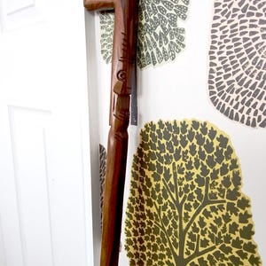 May include: A dark brown wooden walking stick with carved details. The stick has a curved handle and a detailed design on the shaft. The stick is leaning against a white door and wallpaper with a tree pattern.