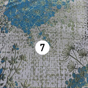 May include: A fabric featuring a light gray background with a woven pattern of bright blue flowers and metallic gold stems and leaves. A white circular icon with the black number 7 is positioned in the center of the floral textile design.