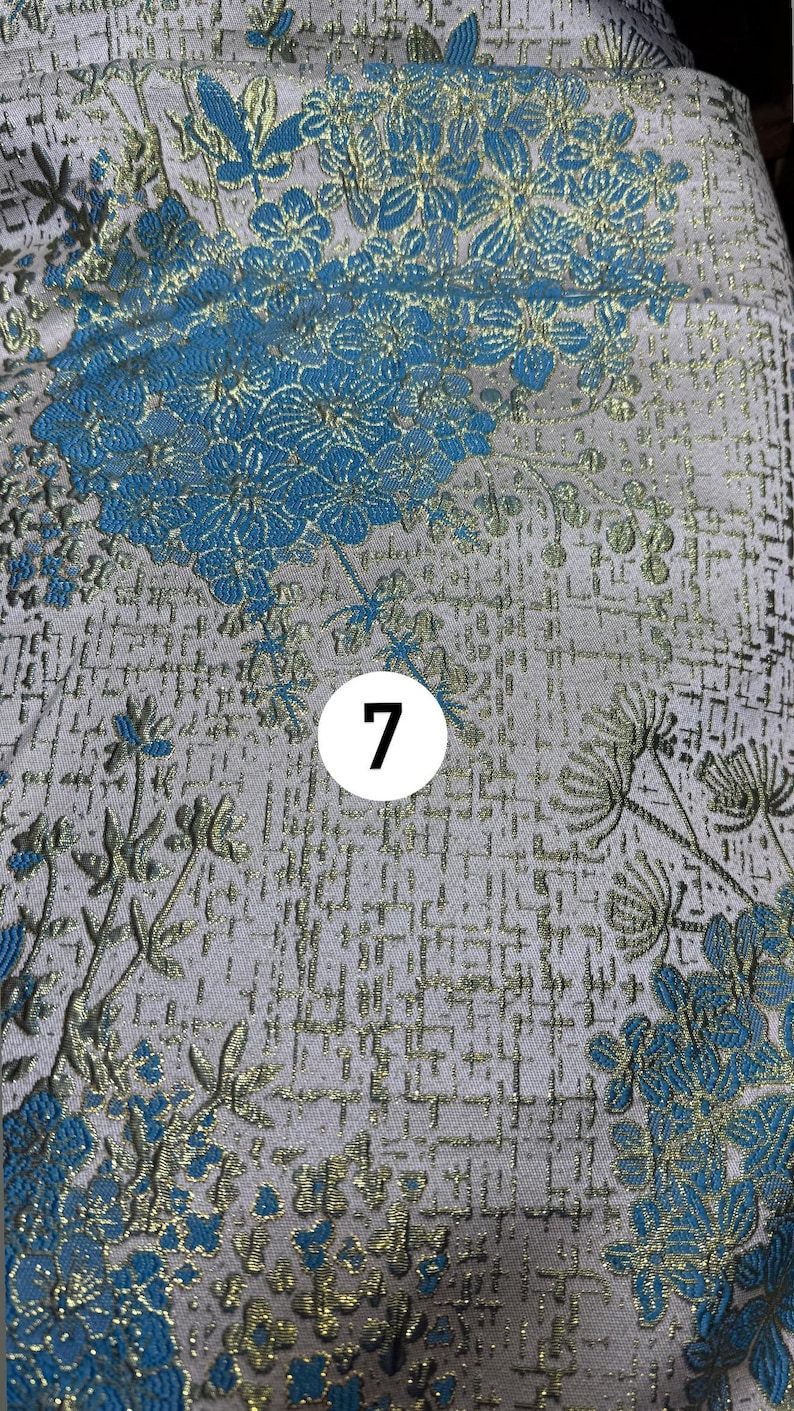May include: A fabric featuring a light gray background with a woven pattern of bright blue flowers and metallic gold stems and leaves. A white circular icon with the black number 7 is positioned in the center of the floral textile design.