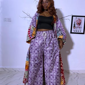 May include: A matching purple and white patterned pants and kimono set. The wide-leg pants have a drawstring waist. The kimono has a colorful patterned design on the sleeves and edges. A black crop top is worn underneath. A framed Vogue magazine cover is visible.