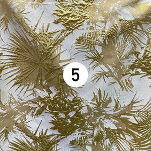 May include: A piece of fabric featuring a white background with a metallic gold palm leaf pattern. The design includes textured, raised gold foliage elements across the surface. A white circular label with the black number 5 is centered.