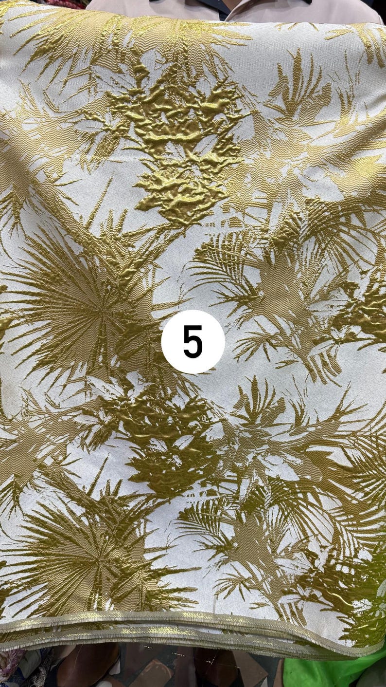 May include: A piece of fabric featuring a white background with a metallic gold palm leaf pattern. The design includes textured, raised gold foliage elements across the surface. A white circular label with the black number 5 is centered.