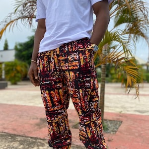 May include: A person wearing a white t-shirt and patterned pants. The pants feature a black, red, and yellow abstract design. The pants have a drawstring waist and are cuffed at the ankles. The person is also wearing a gold watch and sandals.