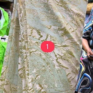 May include: A fabric featuring a light green and white base with a raised, textured gold metallic pattern that creates an abstract, marbled effect across the surface. A red circular icon with the number 1 is positioned in the center.