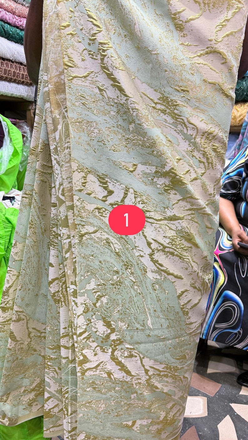 May include: A fabric featuring a light green and white base with a raised, textured gold metallic pattern that creates an abstract, marbled effect across the surface. A red circular icon with the number 1 is positioned in the center.