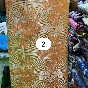 May include: A length of fabric featuring a vibrant orange and gold floral pattern layered over a geometric starburst design. The textile displays a textured, raised surface with a repeating motif and a white circular label with the number 2.
