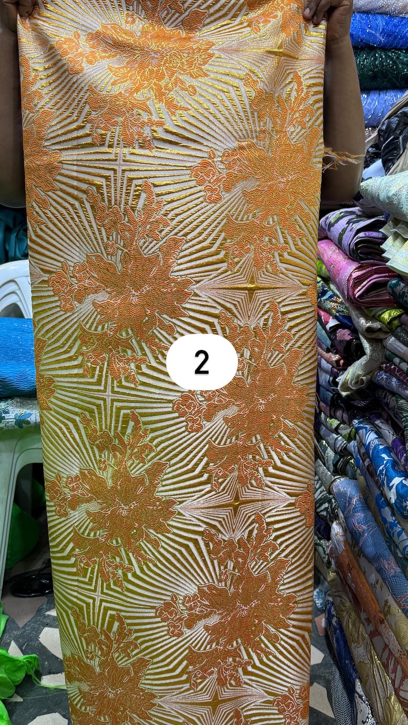 May include: A length of fabric featuring a vibrant orange and gold floral pattern layered over a geometric starburst design. The textile displays a textured, raised surface with a repeating motif and a white circular label with the number 2.