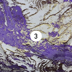 May include: A fabric featuring an abstract pattern in shades of purple, white, and metallic gold. The design consists of flowing, marbled shapes with a textured weave. A white circular label with the black number 3 is centered on the fabric.