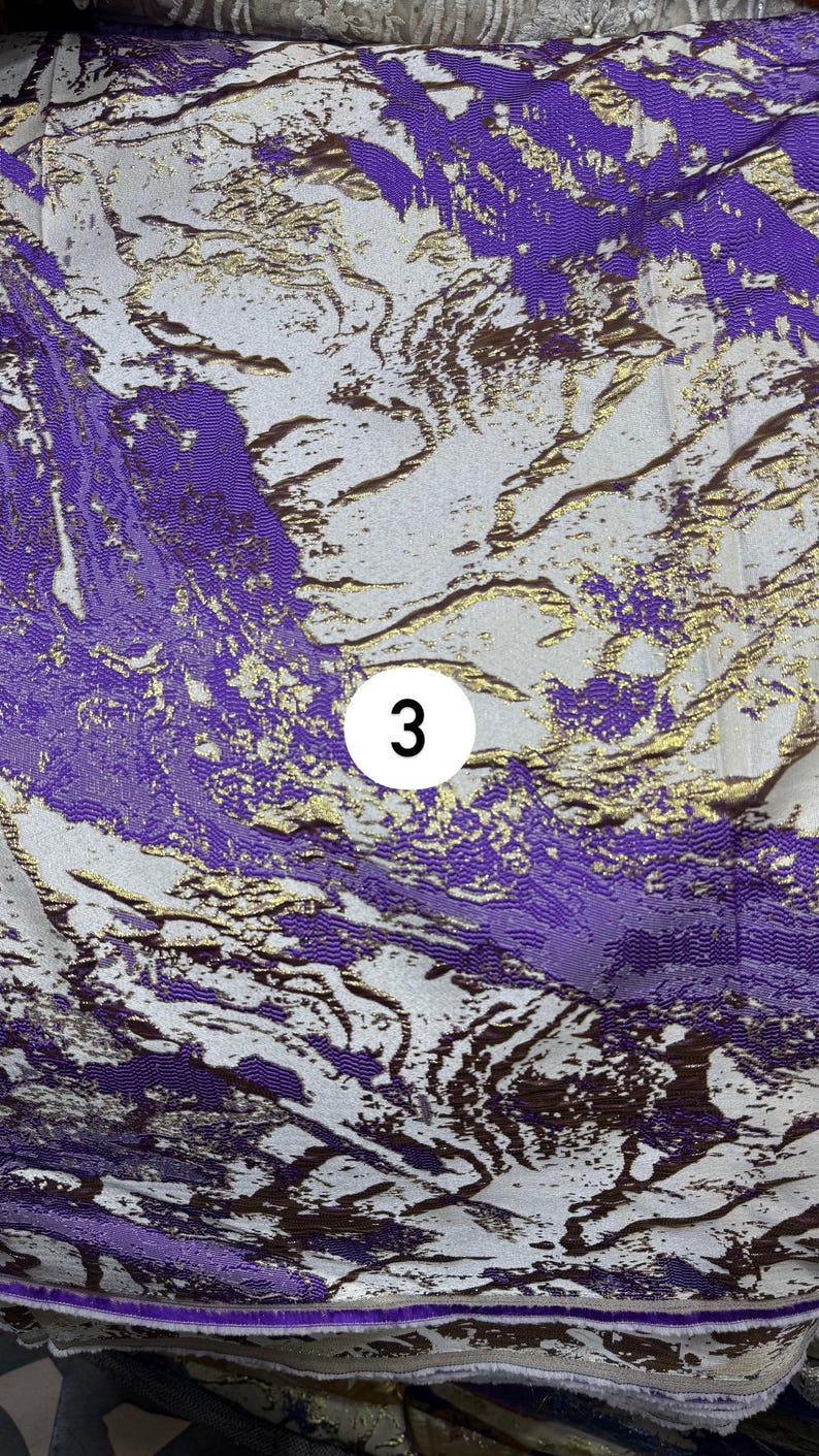 May include: A fabric featuring an abstract pattern in shades of purple, white, and metallic gold. The design consists of flowing, marbled shapes with a textured weave. A white circular label with the black number 3 is centered on the fabric.