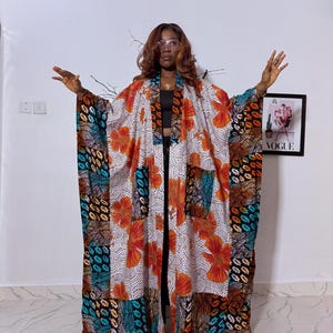 May include: A long, open-front kimono-style robe with a vibrant floral pattern in orange and white. The robe features a patchwork design with sections of patterned fabric in teal, brown, and black. The robe is worn over a black top and pants.