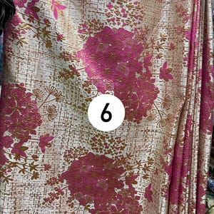 May include: A fabric featuring a cream background with a textured, metallic gold grid pattern and large, vibrant magenta floral clusters. A white circular sticker with the black number 6 is positioned in the center of the textile design.